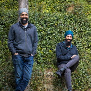 Harpreet and Darsh Singh, two Sikh men in zip up sweatshirts and jeans smiling in front of a wall of green ivy.